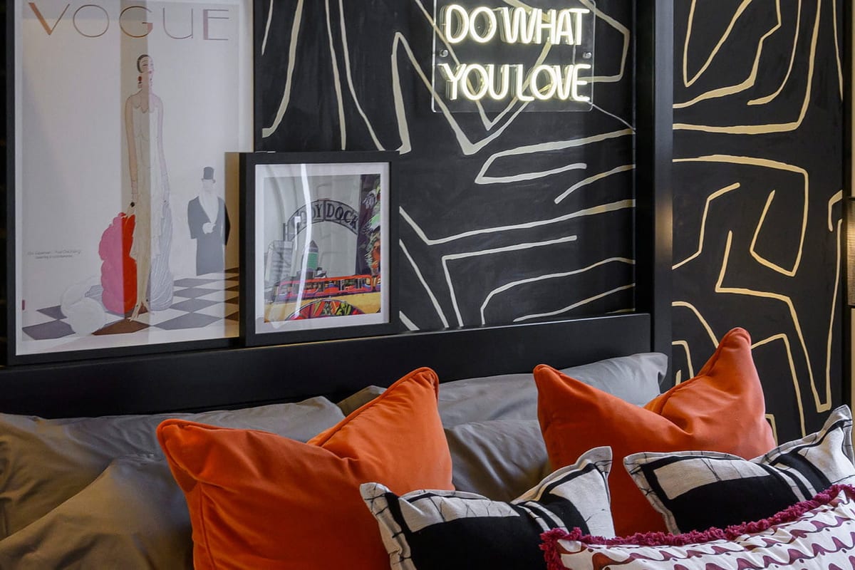 Bedroom with a black and gold graphic feature wall, a neon sign reading Do What You Love and orange velvet cushions