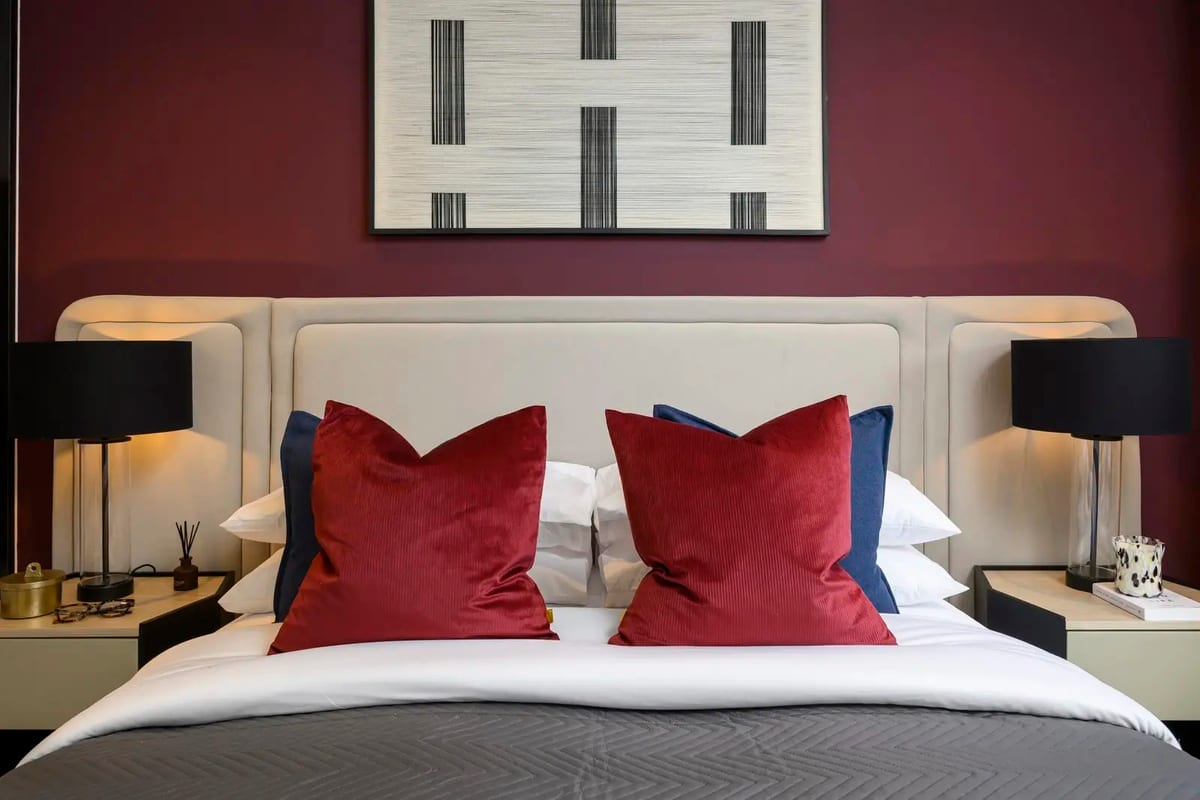Show apartment bedroom seen head-on: a cream upholstered headboard against a deep red wall, red velvet cushions and black bedside lamps