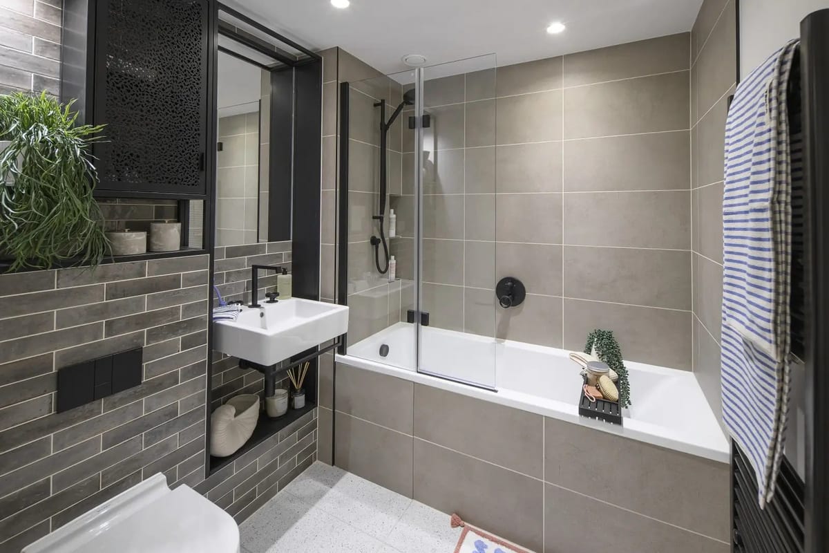 Bathroom with grey glazed tiles, a black-framed mirror cabinet, black brassware and a bath with shower screen
