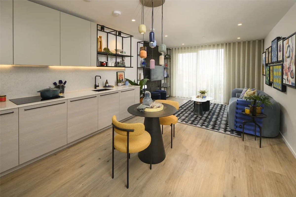The whole open-plan room: a pale kitchen along one wall, a round dining table with mustard chairs, and a blue sofa on a chequered rug by the windows