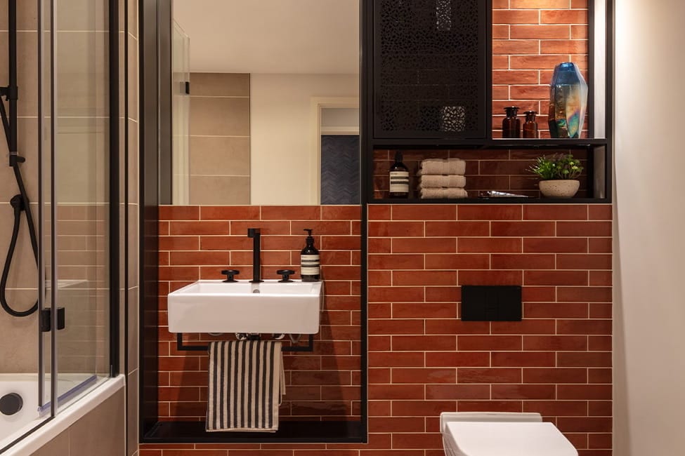 Bathroom with brick-red glazed tiles behind the basin, black fittings, a niche shelf and a bath with shower screen