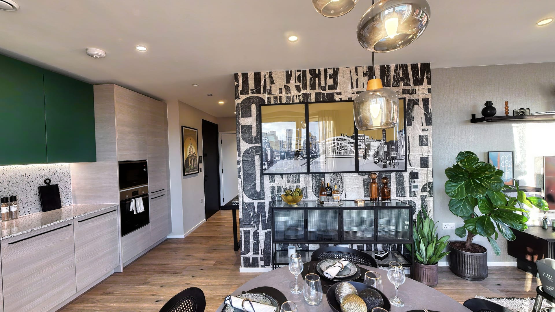 Dining area in The Heron Collection show apartment: a glazed screen wrapped in black-and-white typographic wallpaper, a glass pendant over the table and a forest-green kitchen beyond