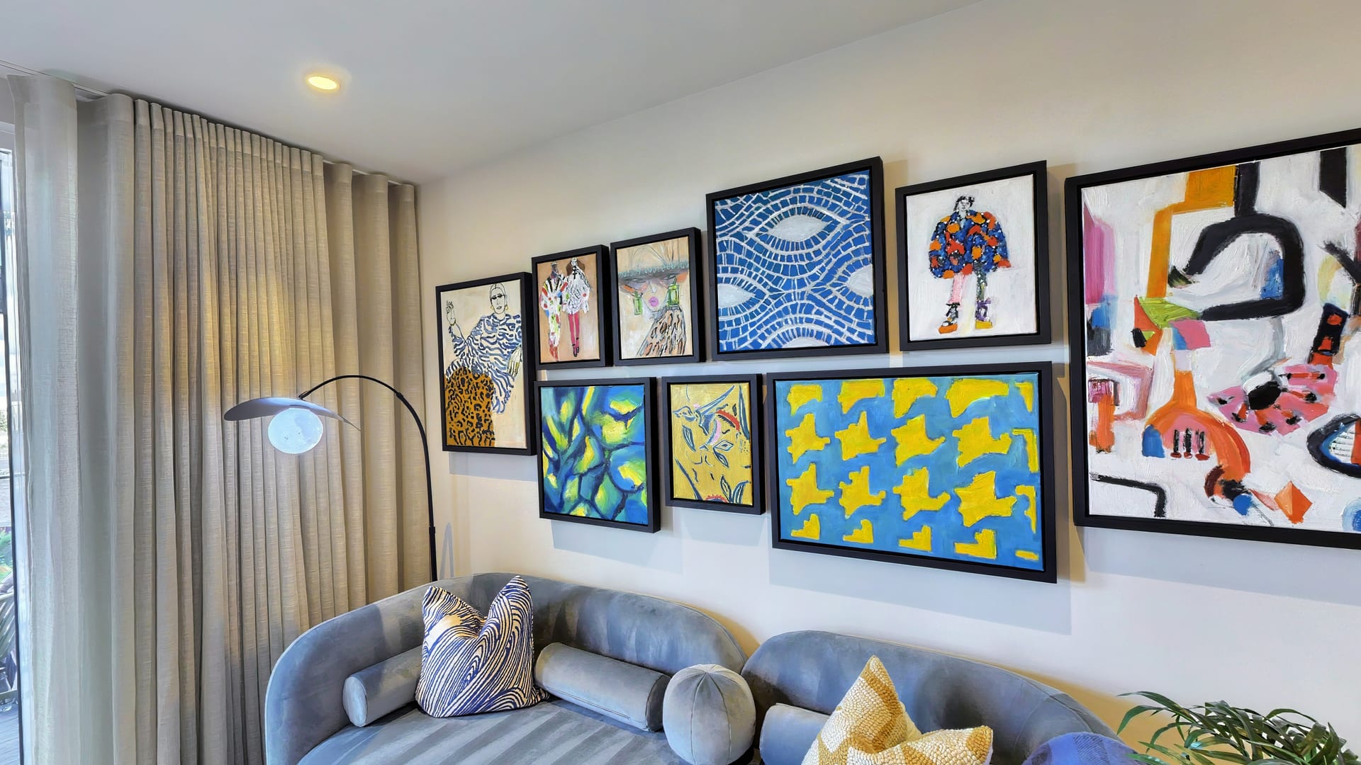 Living room in The Millhouse show apartment: a gallery wall of bold blue, yellow and orange prints above a grey sofa, with an arc floor lamp by full-height curtains