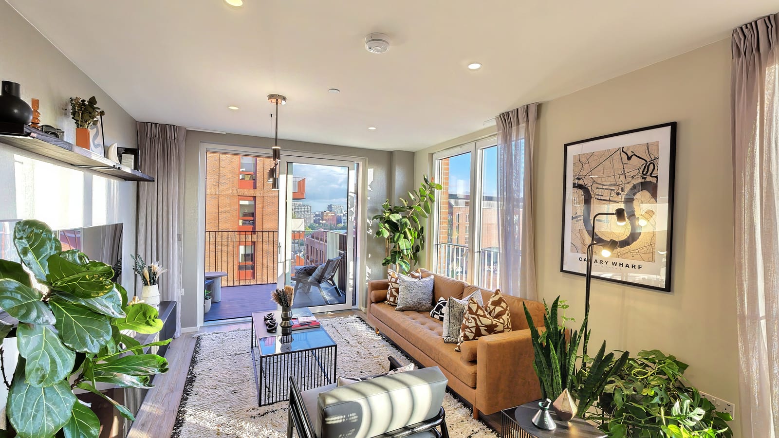 Living room in The Heron Collection show apartment: a tan leather sofa with patterned cushions, plants and a framed print, with the balcony doors and city beyond