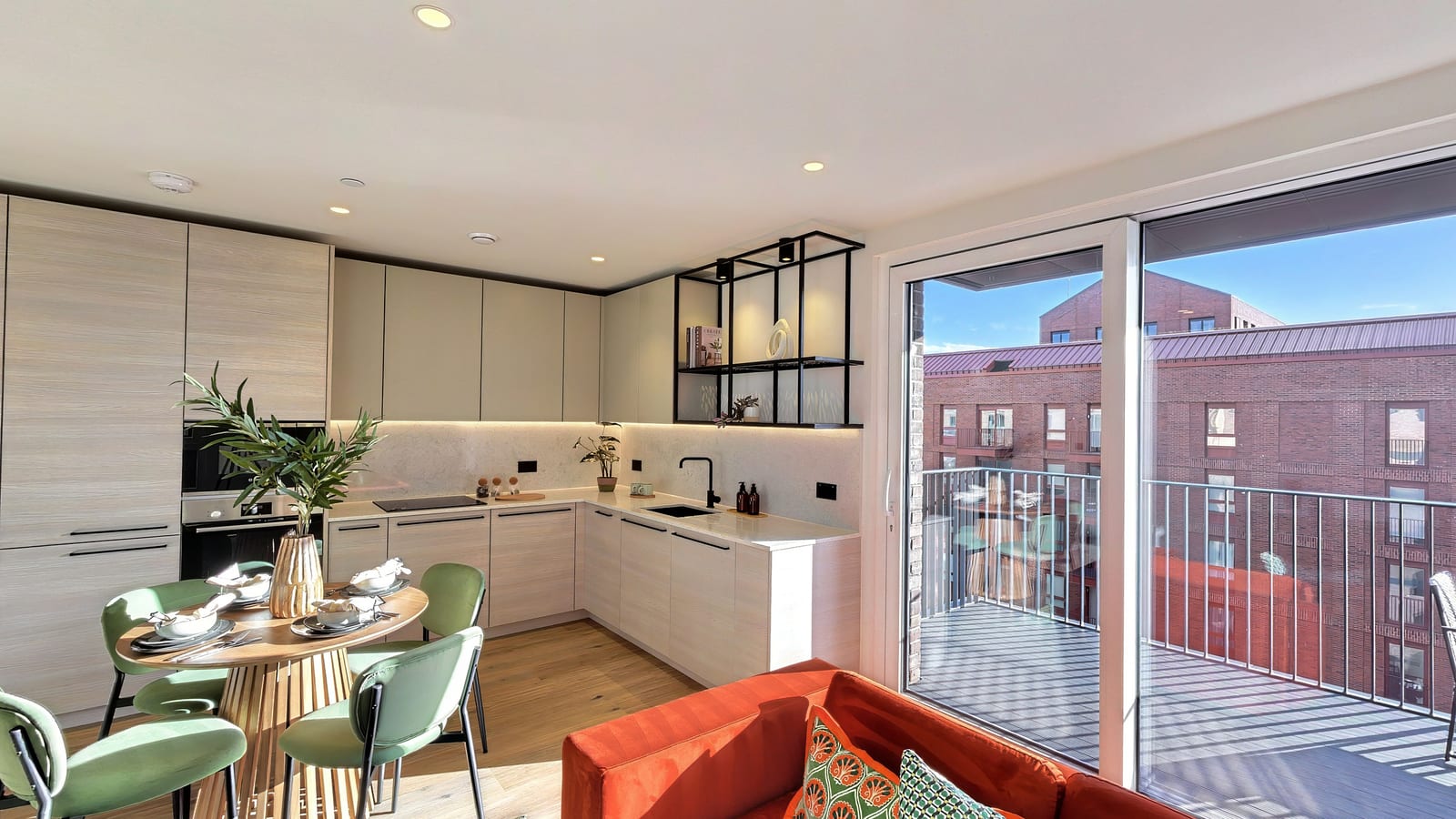 Open-plan room in apartment B1-08-09: a pale kitchen with black open shelving, a round dining table with sage-green chairs, a red velvet sofa and sliding doors to the balcony
